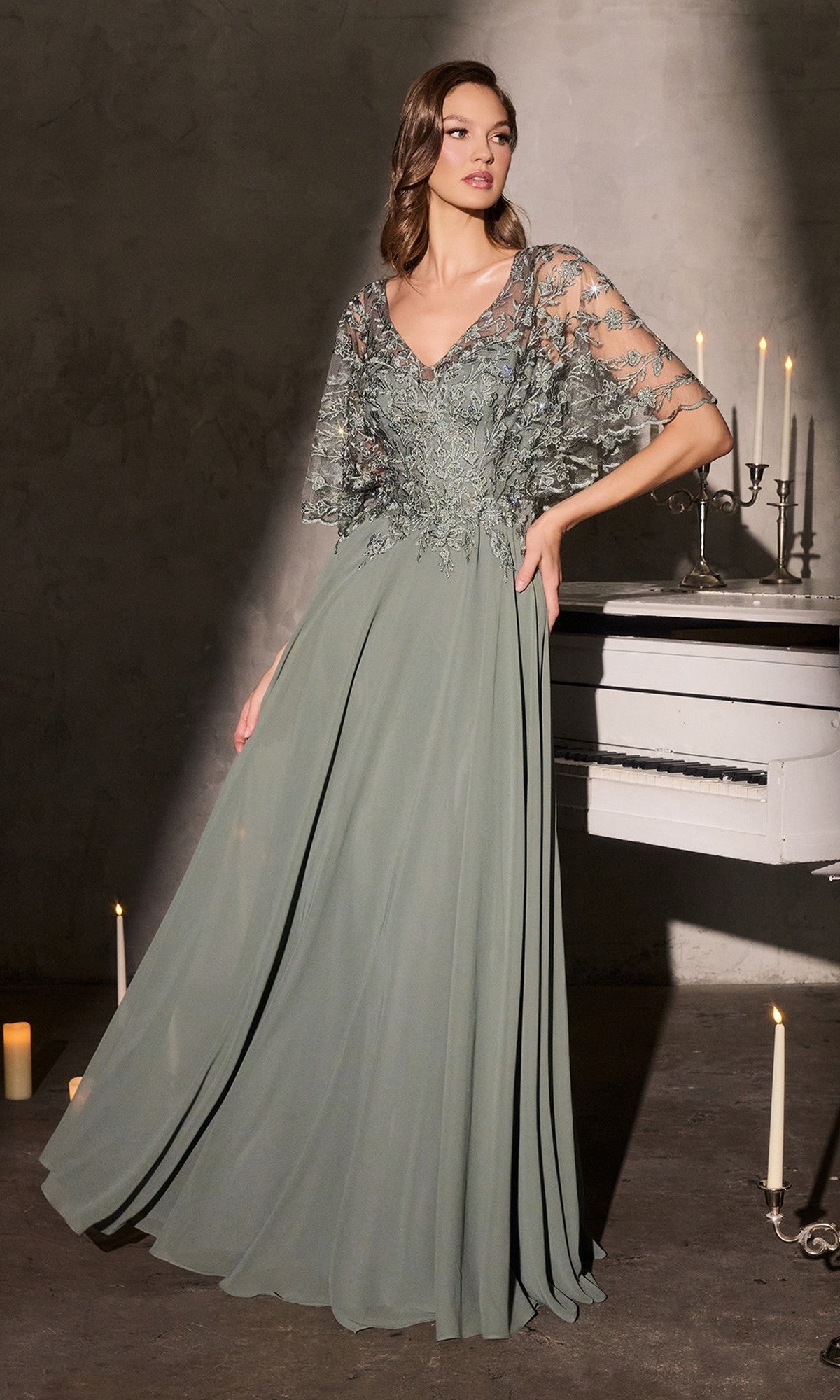 A woman in the Ladivine CL10 long formal dress, moss green with a lace-embroidered bodice, sheer sleeves, and flowing chiffon skirt, stands indoors by a white piano and lit candles.